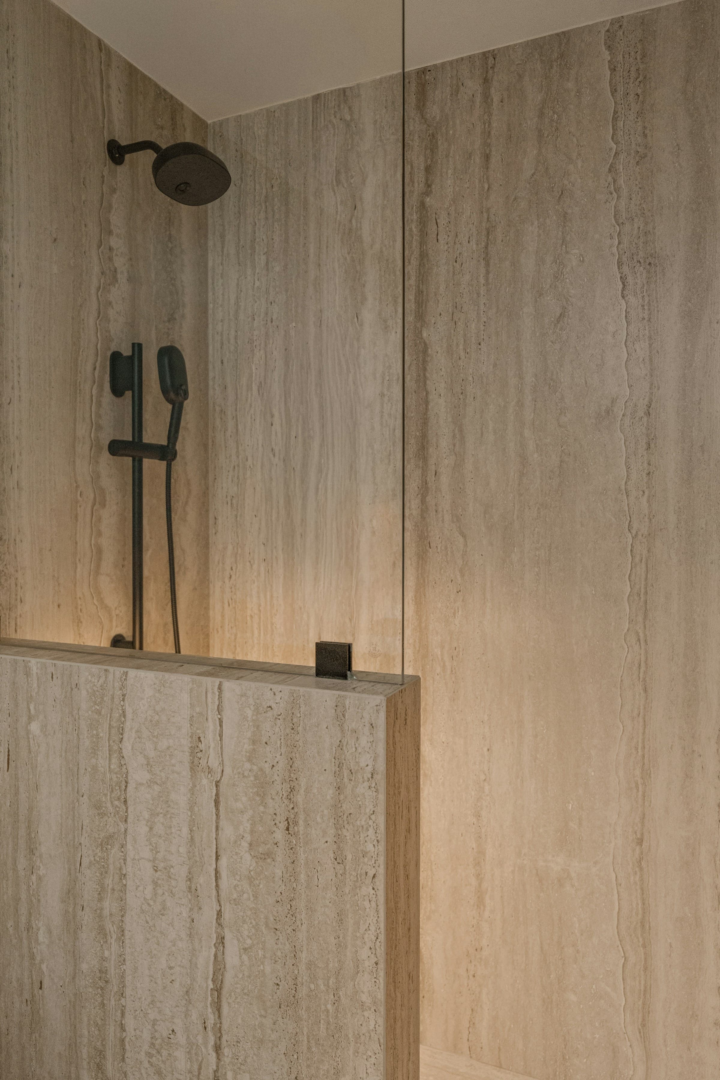 Close-up of a travertine shower wall and half-height partition, its horizontal bedding visible in the stone, with a dark bronze shower head and slide rail.