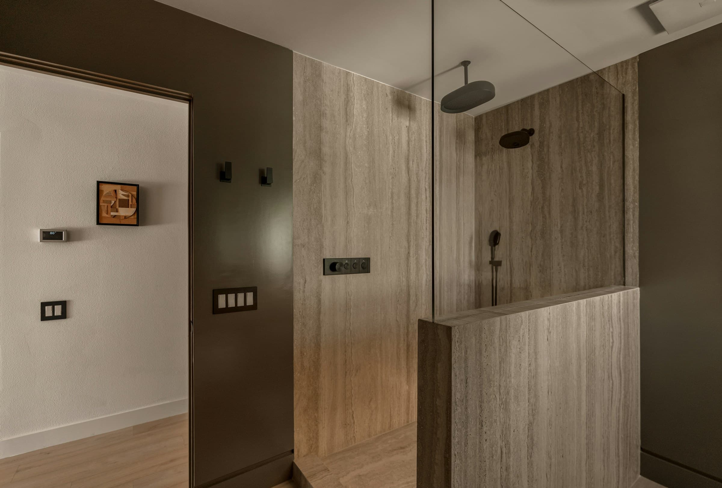 A walk-in shower lined floor to ceiling in travertine, with a frameless glass screen, dark bronze fittings and a deep olive painted wall beyond.