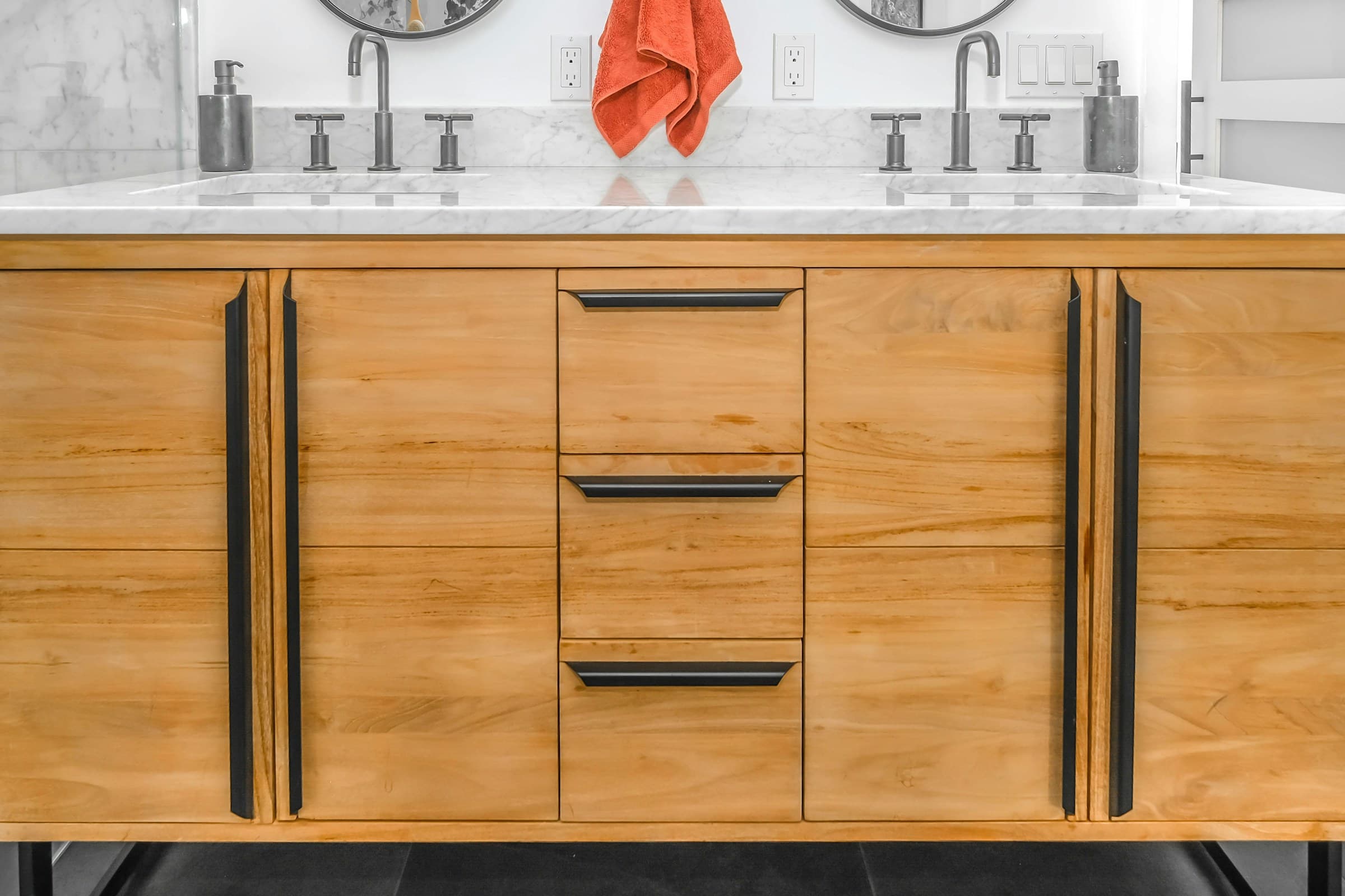 A double bathroom vanity in pale figured wood with integrated dark pulls, a marble countertop with two undermount basins, cross-handle faucets and round mirrors above.