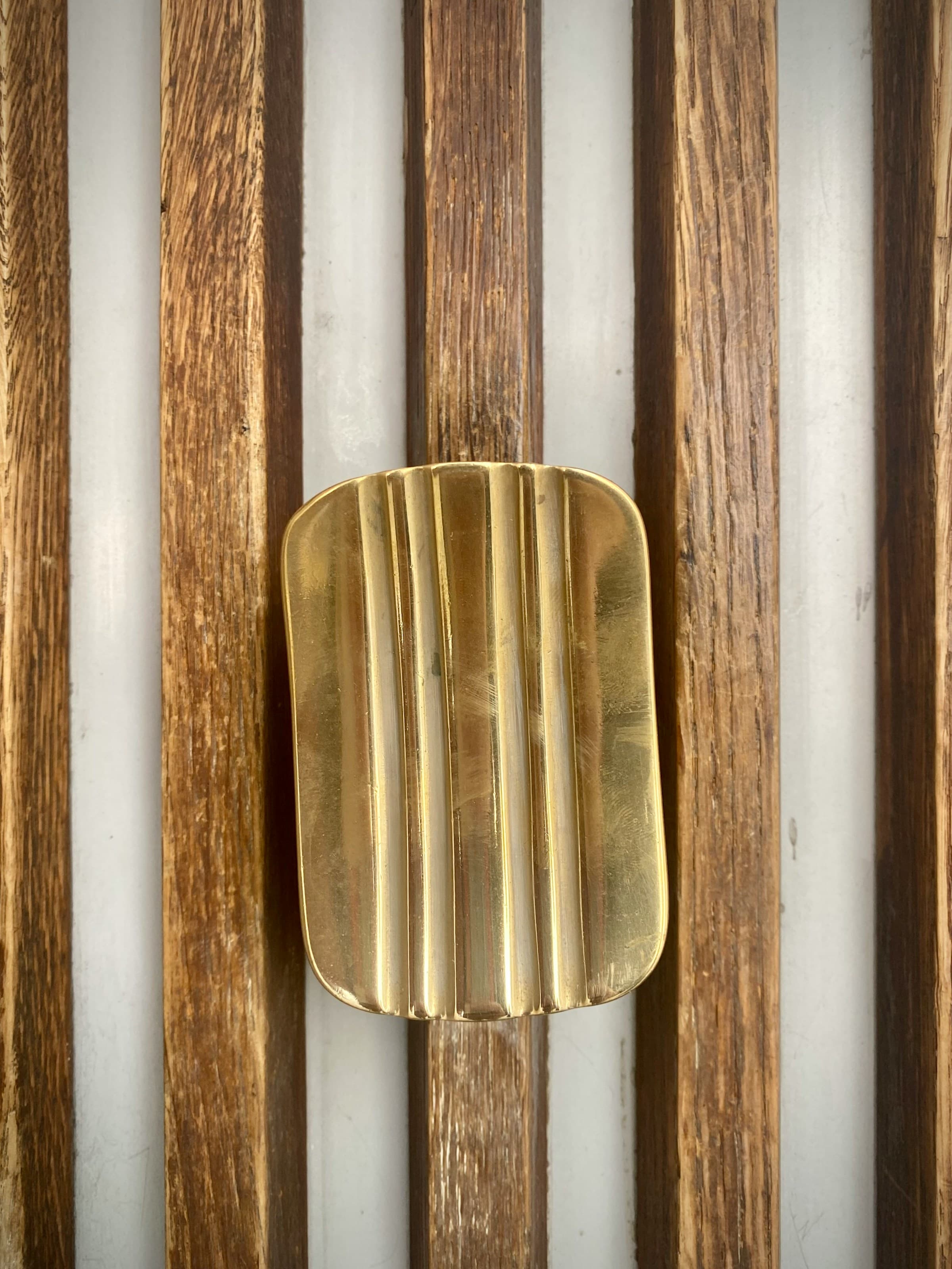 Close-up of a fluted unlacquered brass handle mounted on a wall of narrow oak slats, the brass polished bright where hands have touched it and darker in the flutes.