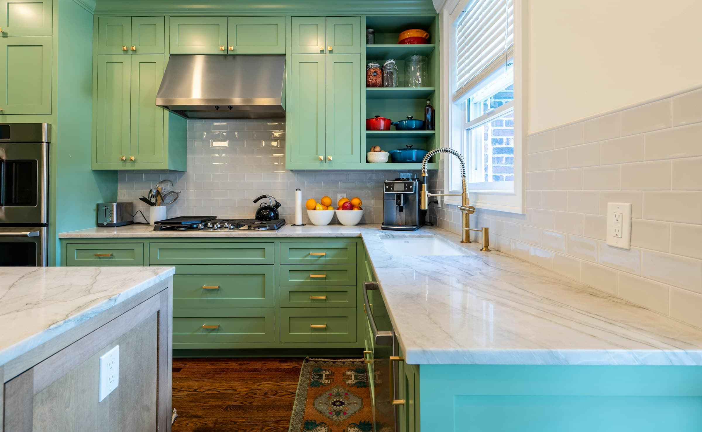 A green painted kitchen with shaker cabinets, brass pulls, a marble counter and backsplash, open shelving holding cookware, and daylight from a window over the sink.