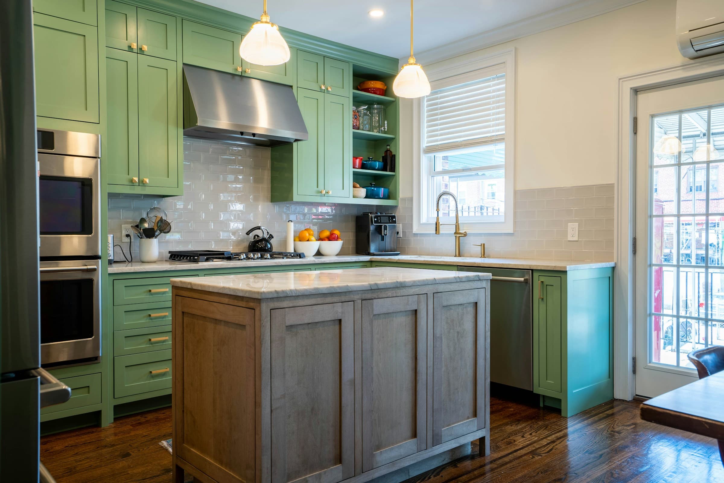 A kitchen with green cabinetry, a stainless range hood, two glass pendant lights over a wood island topped in marble, and a door to a balcony letting in daylight.
