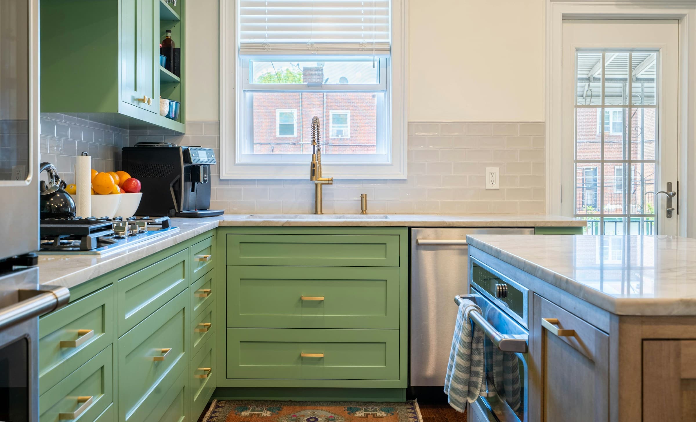 A green kitchen seen along the counter, with a gas range under a stainless hood, open shelving stacked with pots, marble counters and a window over the sink.