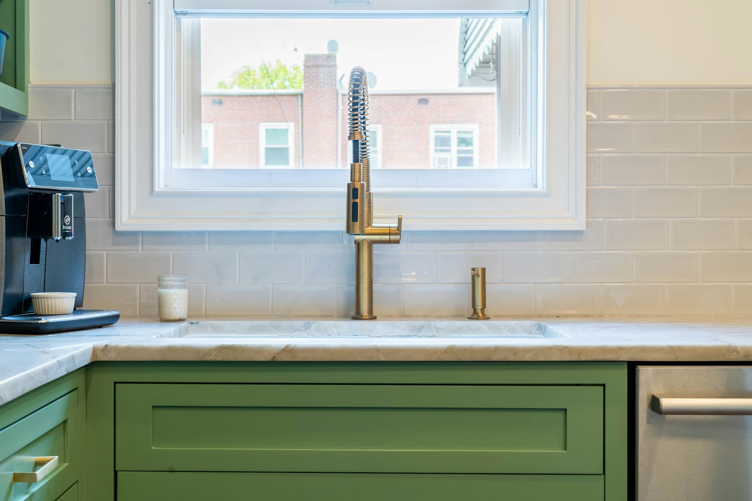 A kitchen sink set into a marble counter beneath a window, with an unlacquered brass faucet, green painted cabinet fronts below and a pale glazed tile backsplash catching the daylight.