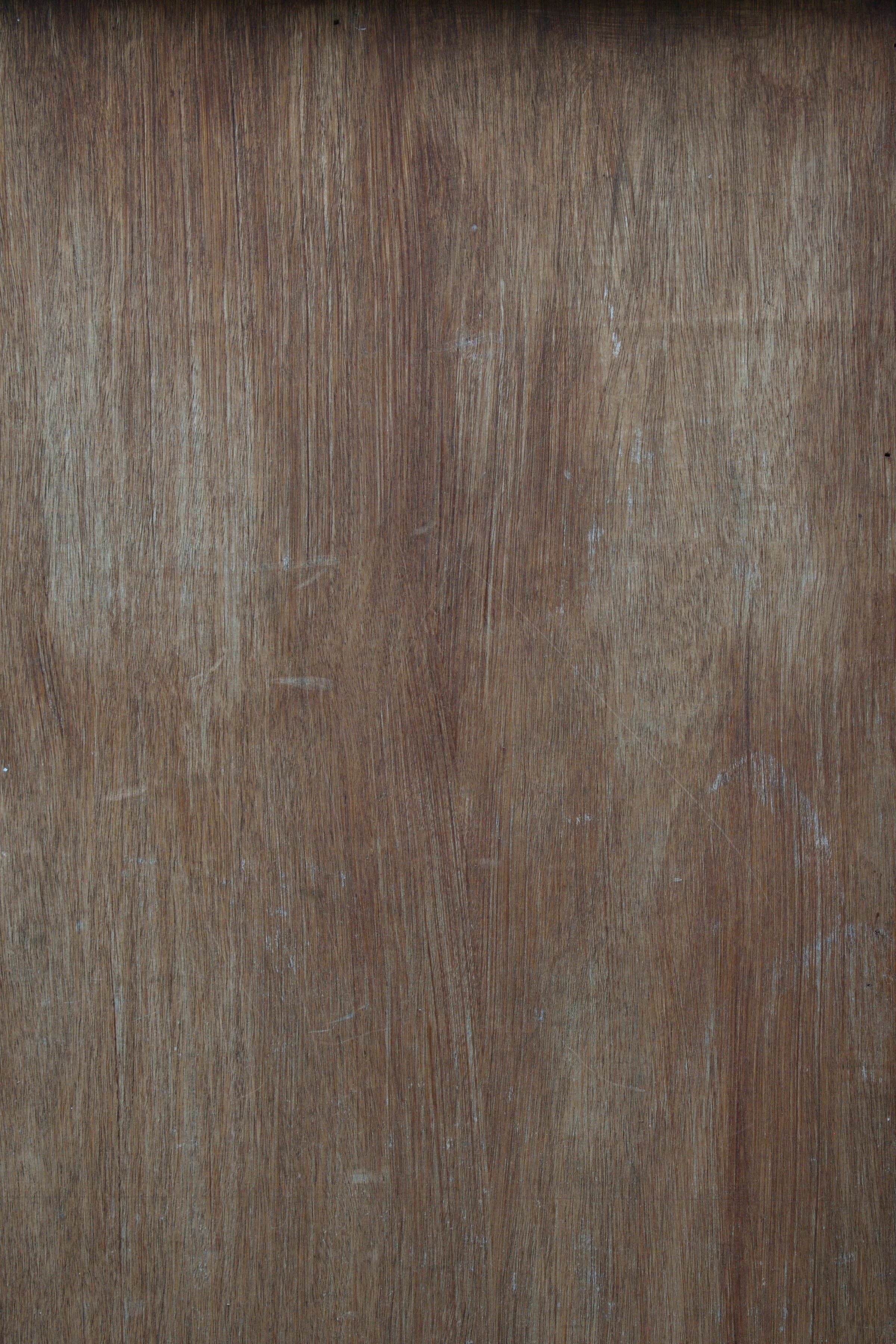 A flat panel of brown hardwood filling the frame, its straight grain running vertically, the surface marked with fine pale scratches and small scuffs.