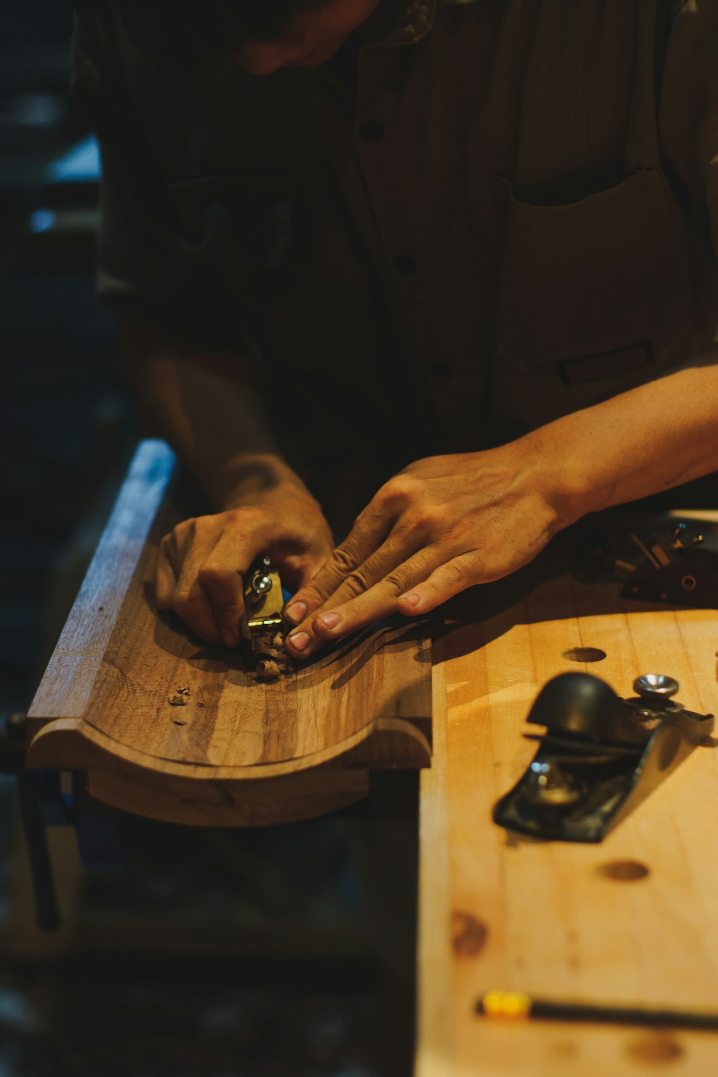 Two hands working a small brass block plane along the curved edge of a hardwood panel clamped to a bench, shavings gathered on the surface and larger planes resting alongside.