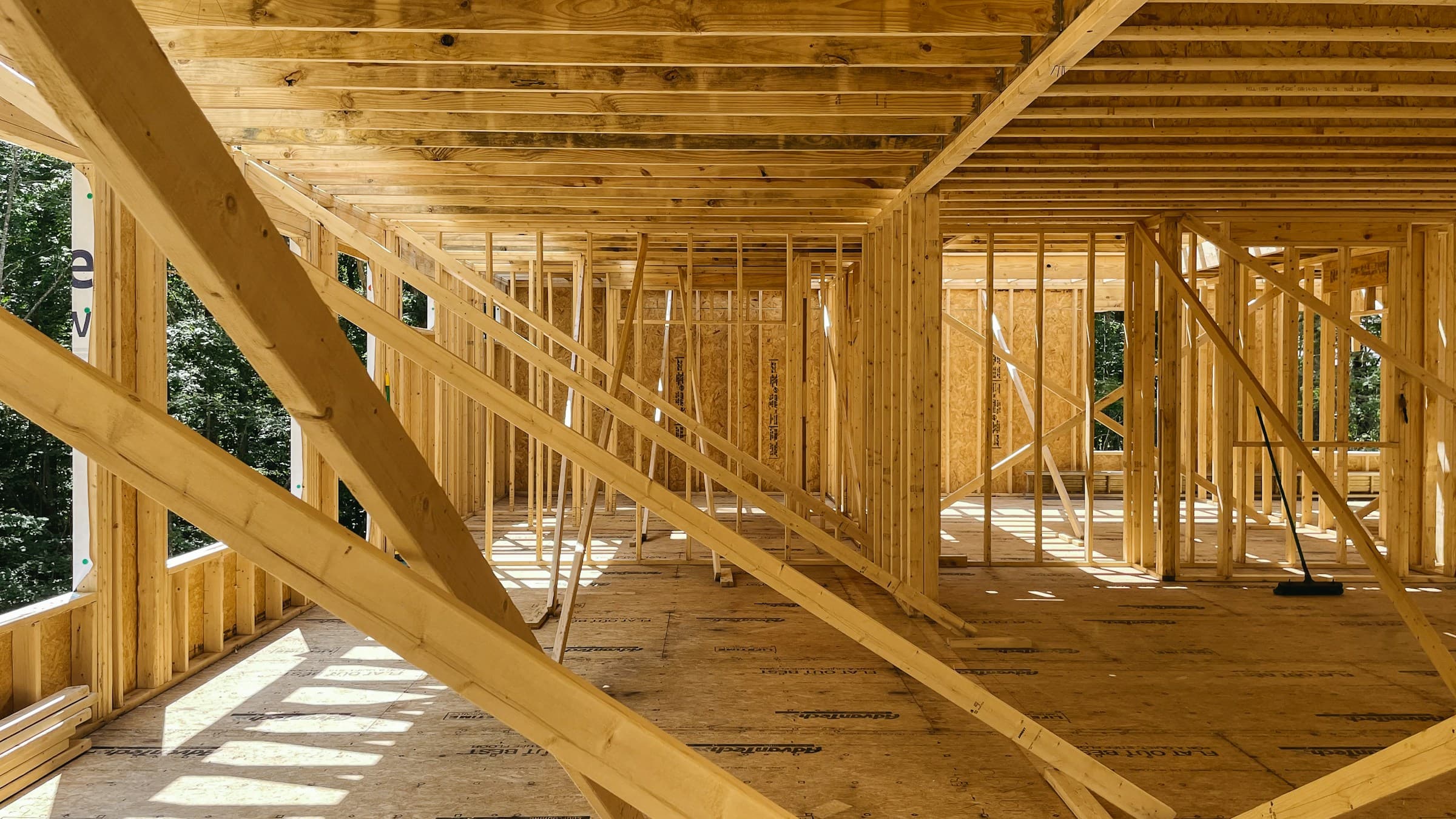 The inside of a timber-framed addition before sheathing, with floor joists overhead, braced stud walls, sheathing panels and sunlight falling across the subfloor.