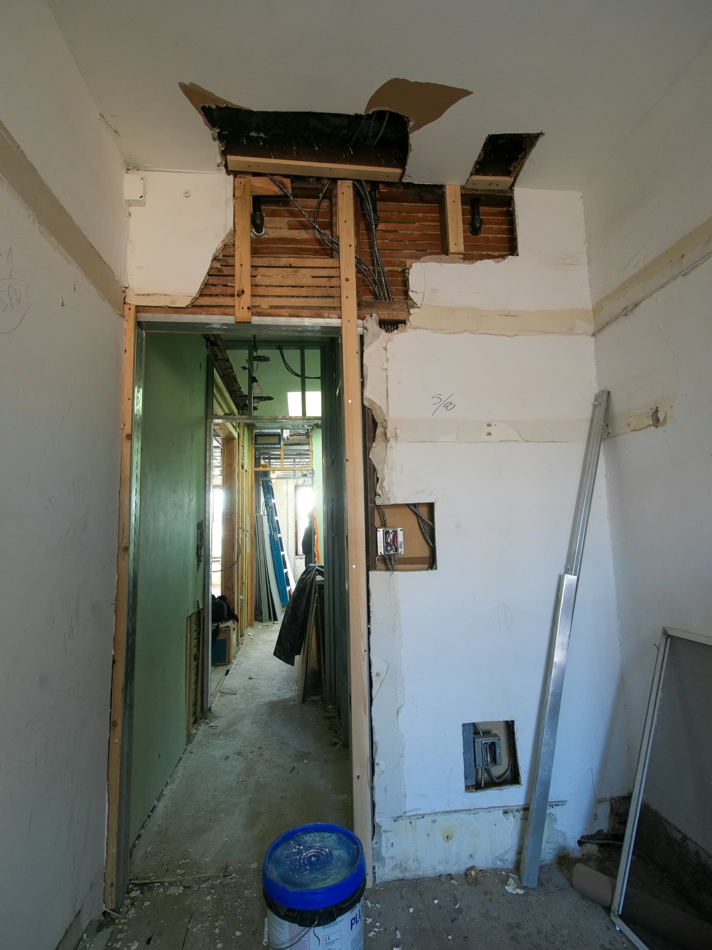A doorway during renovation with the plaster removed above it, exposing horizontal timber lath and old brick, new studs framing the opening, and armored cable dropping through the ceiling.