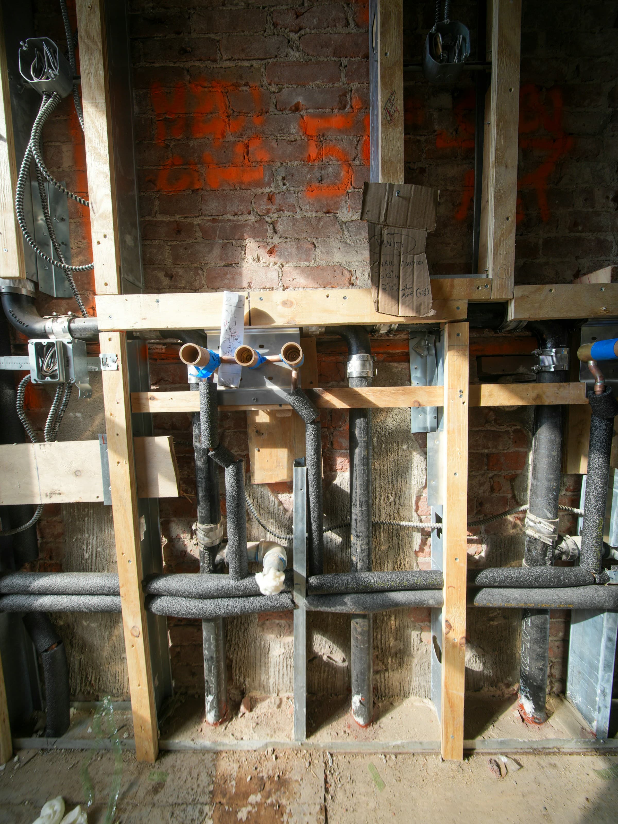 Plumbing rough-in against an old brick wall: copper stubs capped in blue, insulated waste pipes running down between new timber studs, armored electrical cable, and orange survey marks sprayed on the brick.