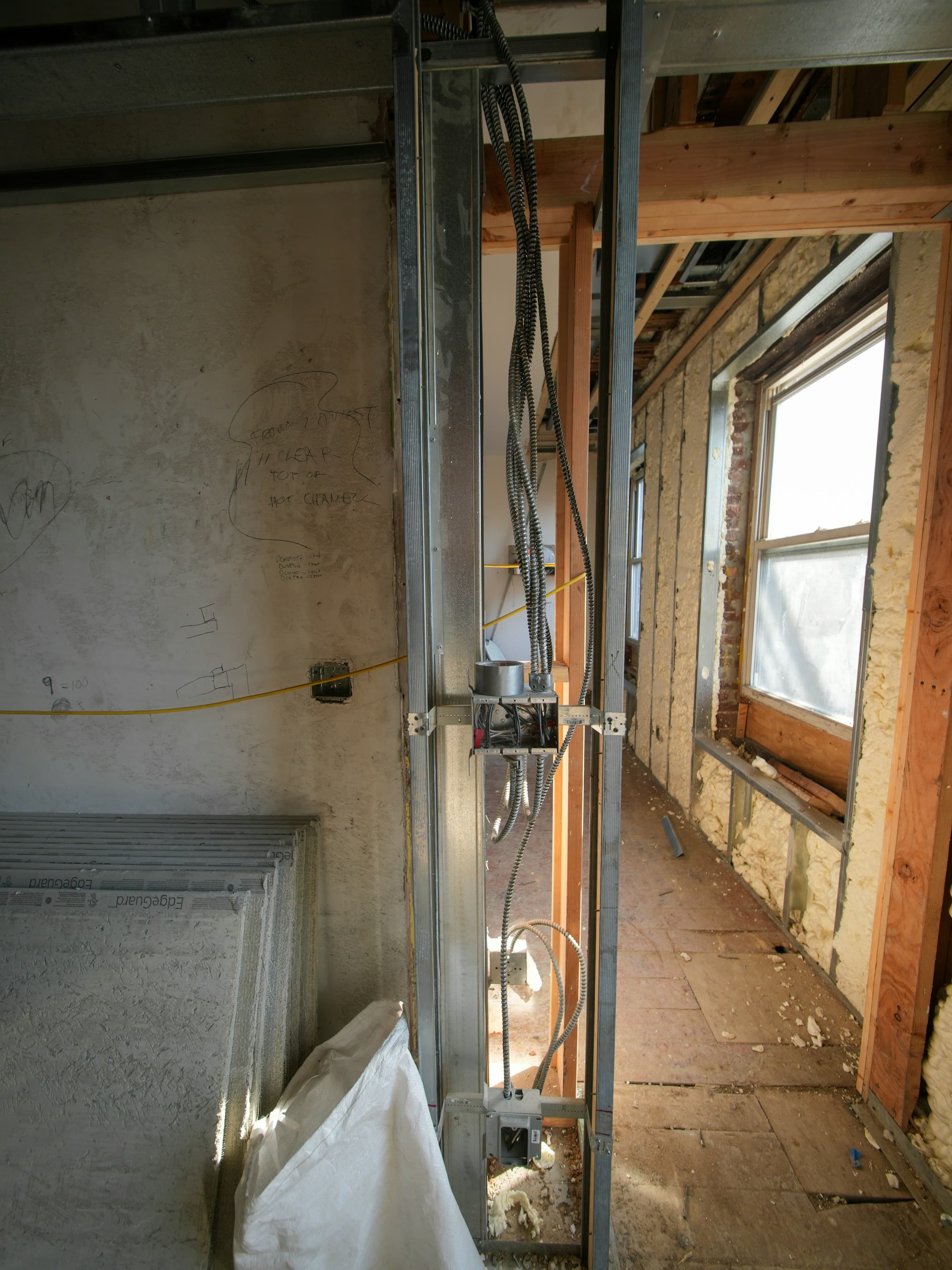 A room stripped back during renovation, with metal studs, armored cable looped down to an open junction box, spray foam insulation around a window, stacked drywall, and contractor notes written on the old plaster.