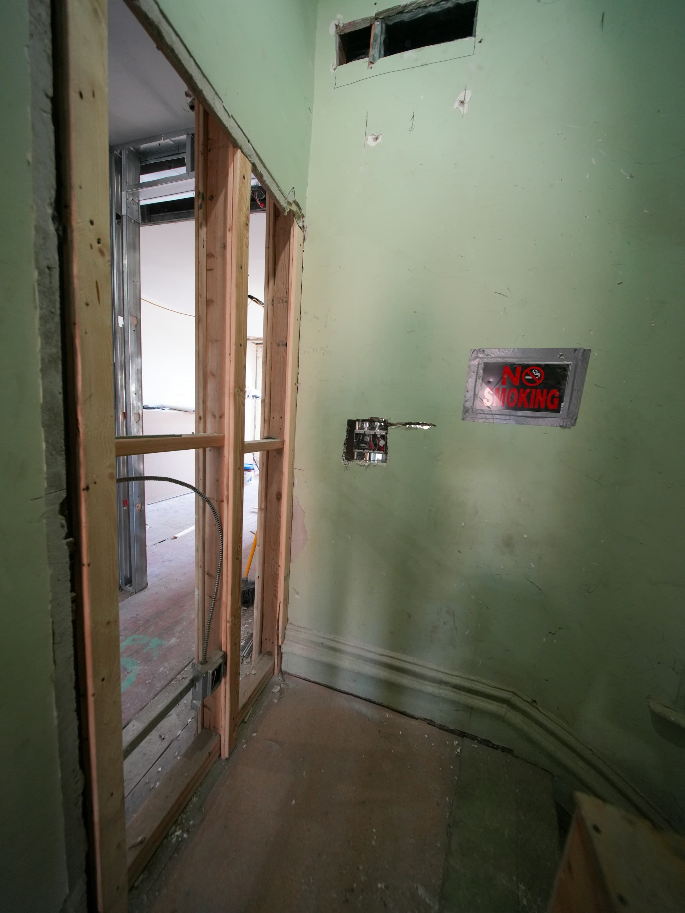 An interior corner during demolition: a stud wall opened up beside a pale green plastered wall, an electrical box hacked out of the plaster, a taped no-smoking sign, and a date pencilled on the wall.